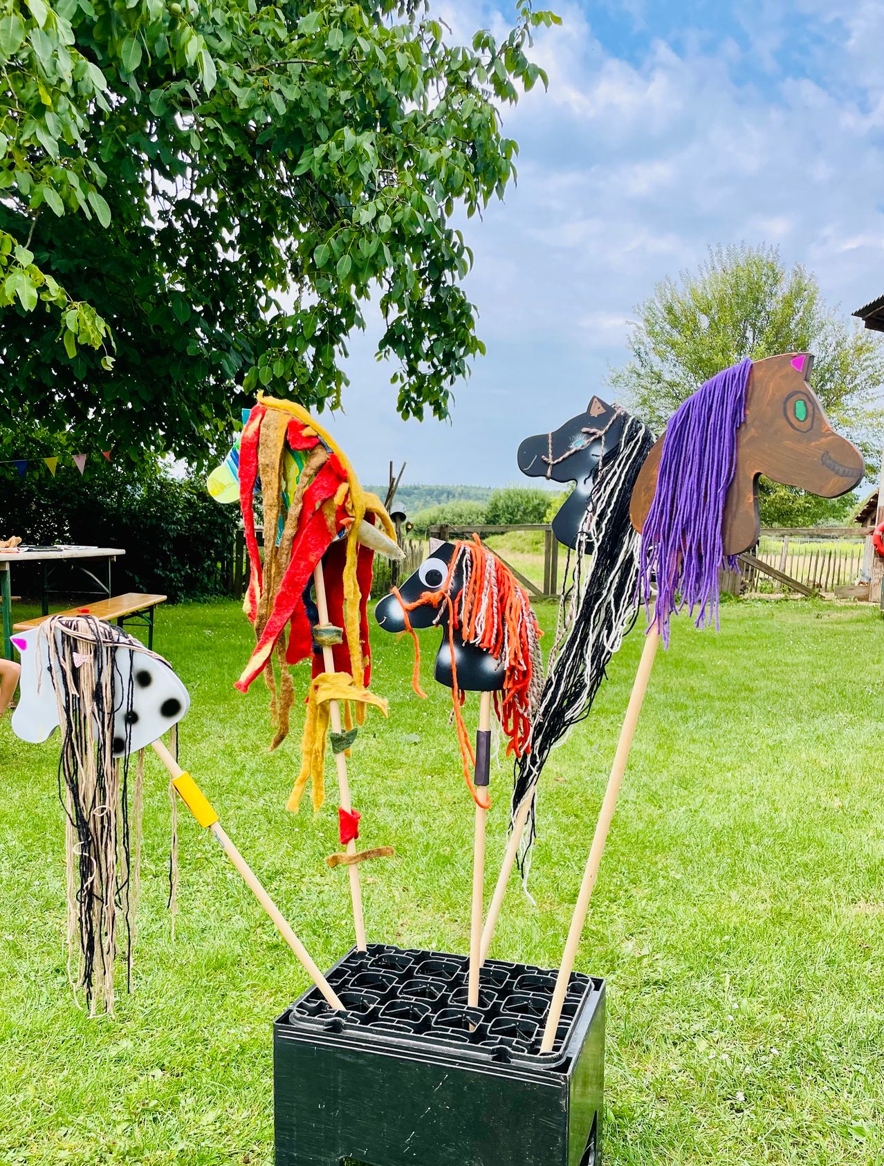 Fünf hölzerne Spielzeugpferde mit bunten Mähnen und Schwänzen sind auf einer Rasenfläche ausgestellt. Die Pferde stehen auf einem schwarzen Teppich mit einem Labyrinthmuster. Dahinter befinden sich ein Holzzaun, Bäume und ein Gebäude.