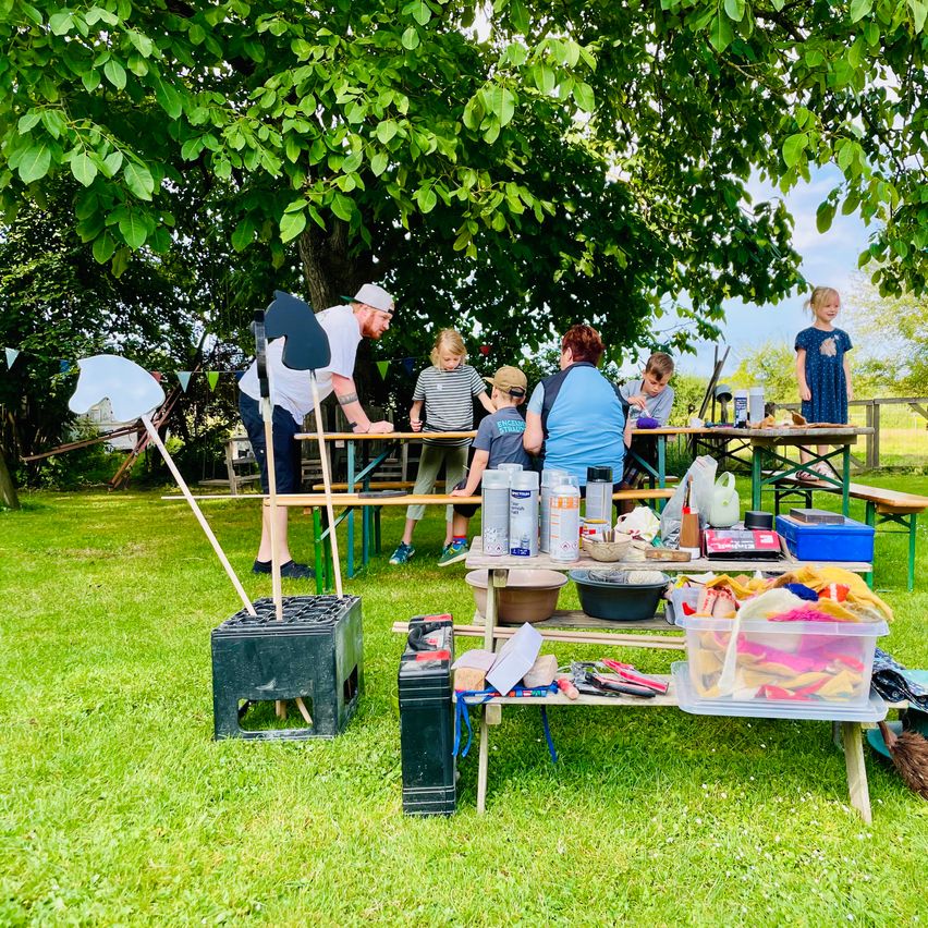 Eine Gruppe von Menschen, einschließlich Kindern, versammelt sich unter einem Baum in einem Park und beschäftigt sich mit verschiedenen Aktivitäten. Ein Mann steht in der Nähe eines Tisches mit Werkzeugen und Materialien.