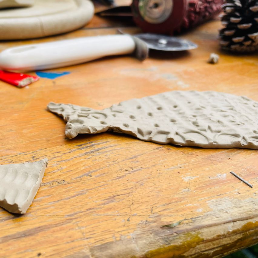 Ein Holztisch mit einem fischförmigen Tonstück, einem Tonwalzen, einem Messer und einem Tannenzapfen.