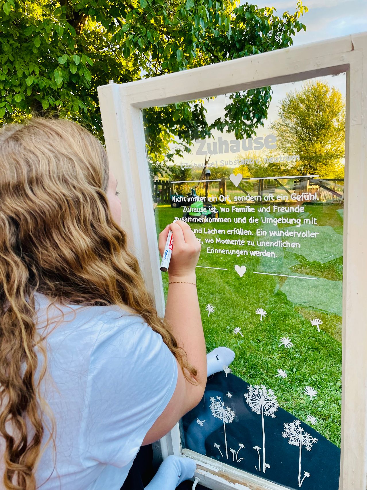 Ein junges Mädchen schreibt mit einem Marker auf eine Glastafel, die Tafel hat einen deutschen Text, der 'Zuhause' und ein Herzsymbol zeigt. Die Tafel ist in Weiß gerahmt und reflektiert die Umgebung, einschließlich eines Grasfeldes und Bäume.