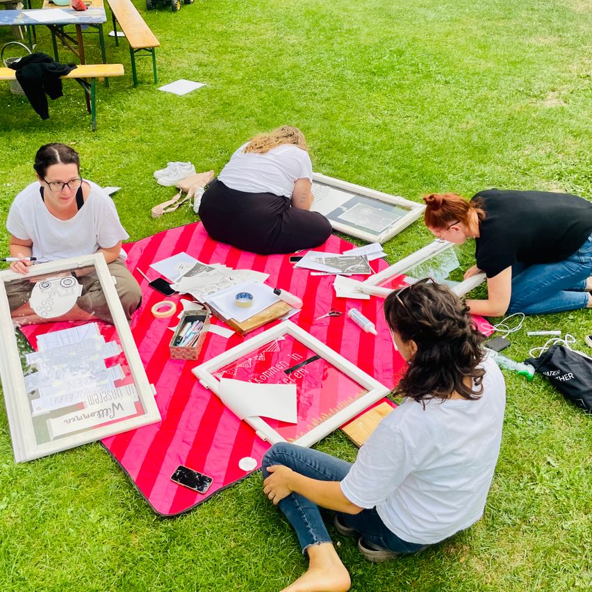 Vier Frauen arbeiten in einem Park auf einer roten Decke an einem Projekt. Sie sind von Papieren, Rahmen und Kunstzubehör umgeben. Eine Frau hält einen Marker.