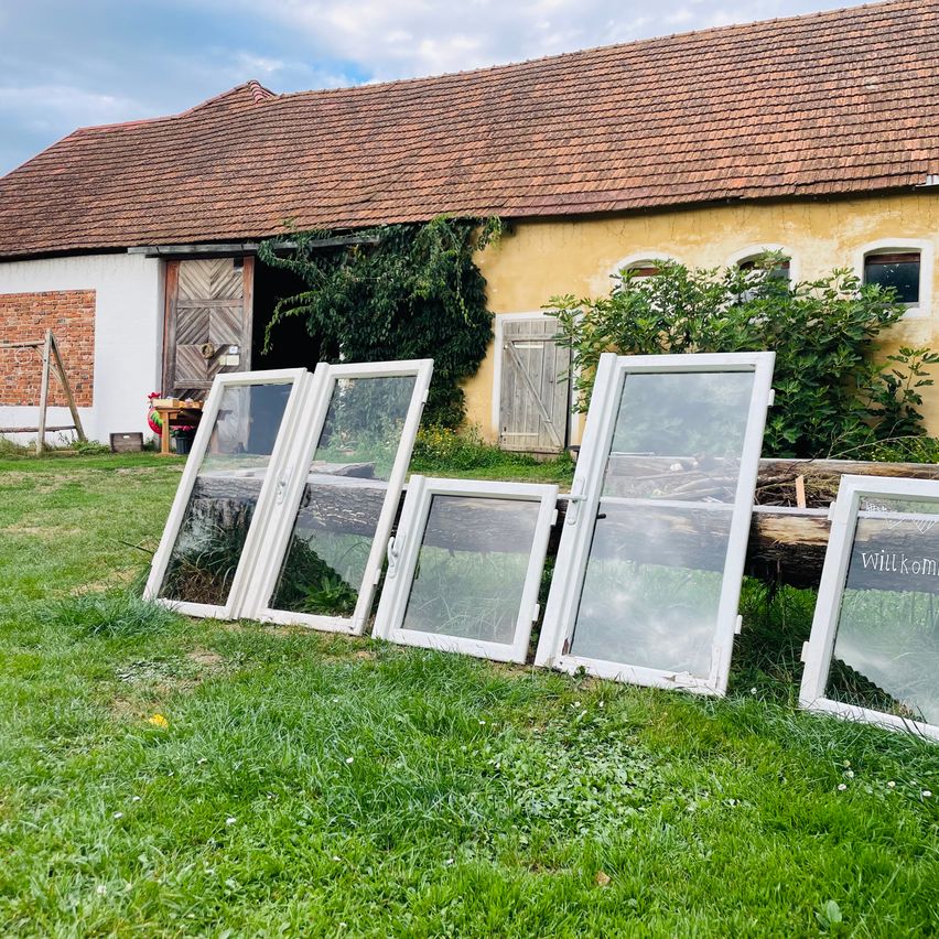 Fünf alte weißgerahmte Fenster sind auf einem Rasen vor einem gelben Gebäude mit einem braunen Dach und einer Holztür angeordnet.