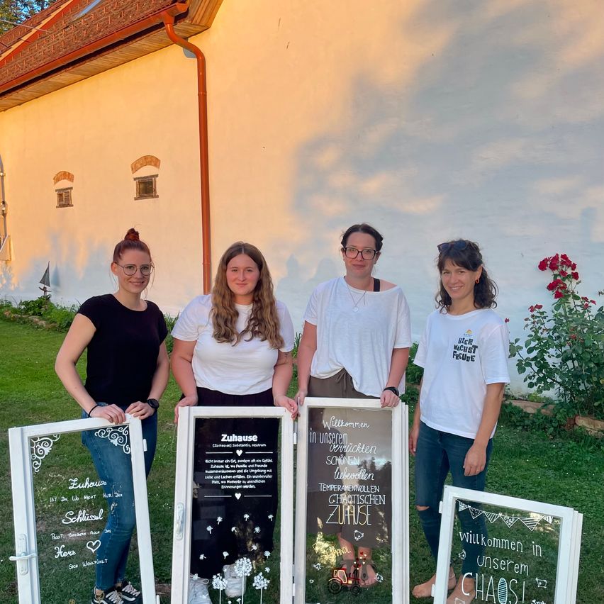 Vier Frauen stehen vor einem Gebäude, jede hält eine Glastür mit dekorativer Beschriftung. Die Türen haben florale und verspielte Designs. Die Frau auf der rechten Seite trägt ein weißes T-Shirt mit den Worten 'Duer Muechst Fude'. Dahinter steht ein weißes Gebäude mit rotem Rand und einem Garten mit roten Blumen.