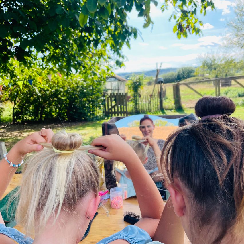 Ein Mädchen mit blonden Haaren lässt sich von einem anderen Mädchen im Garten die Haare stylen, mit Blick auf ein Feld und Bäume.
