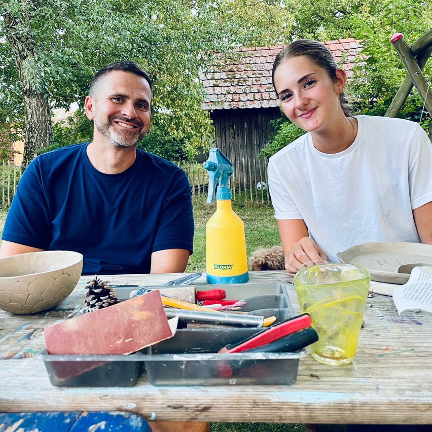 Ein Mann und eine Frau sitzen lächelnd an einem Tisch im Freien, umgeben von Töpferwerkzeugen und -materialien. Der Mann trägt ein blaues Shirt, die Frau ein weißes. Hinter ihnen steht ein Holzhaus und Bäume.