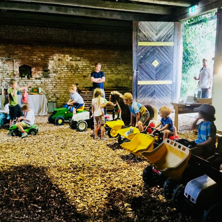 Eine Gruppe von Kindern spielt mit Spielzeugtraktoren in einer Scheune mit einer Ziegelmauer und einer Holztür.