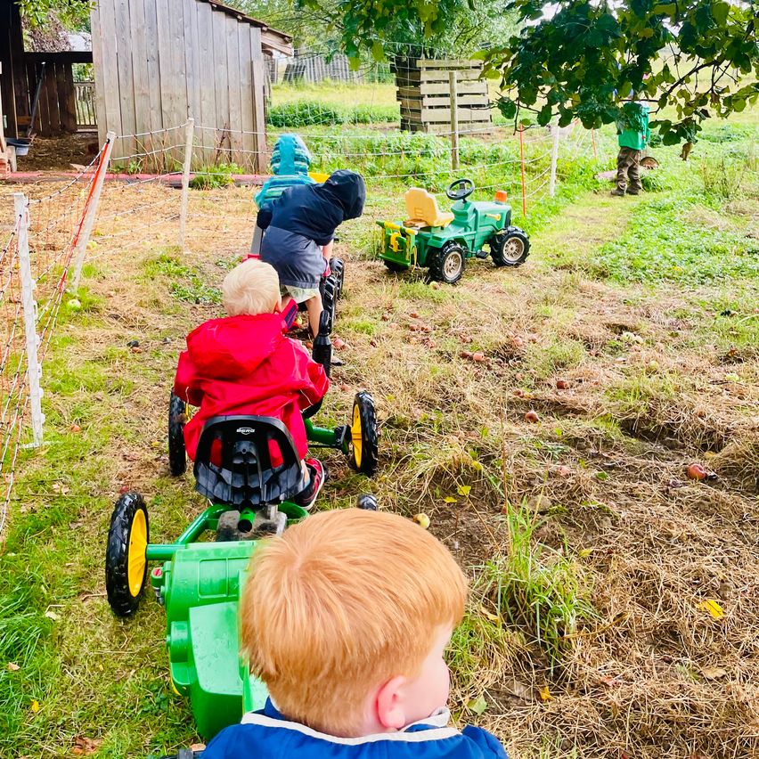 Vier Kinder in Regenmänteln fahren mit Spielzeugtraktoren auf einem Grasfeld, im Hintergrund ein Bauernhaus.