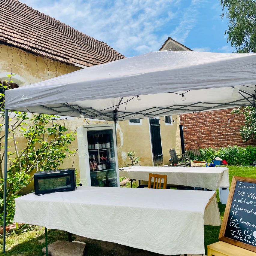 Ein Outdoor-Essenstand unter einem Zelt mit einem Mini-Kühlschrank und einer Tafel mit deutschem Text. Der Stand ist in einem Garten mit tischdeckenbedeckten Tischen aufgestellt.