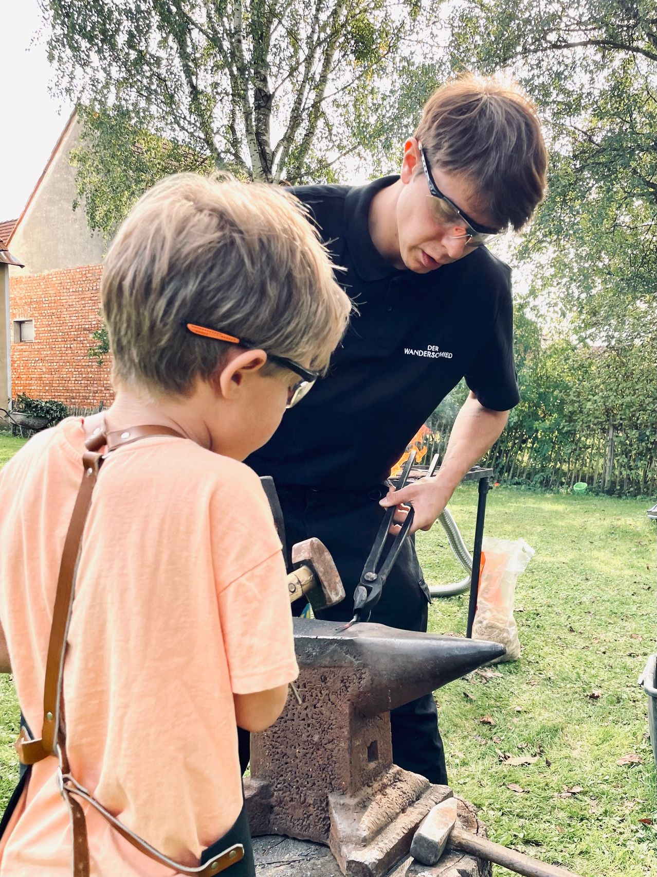 Ein junger Junge lernt von einem Mann mit Brille und schwarzem Hemd, wie man einen Hammer und Amboss benutzt. Sie sind draußen in der Nähe eines Hauses und eines Rasens.