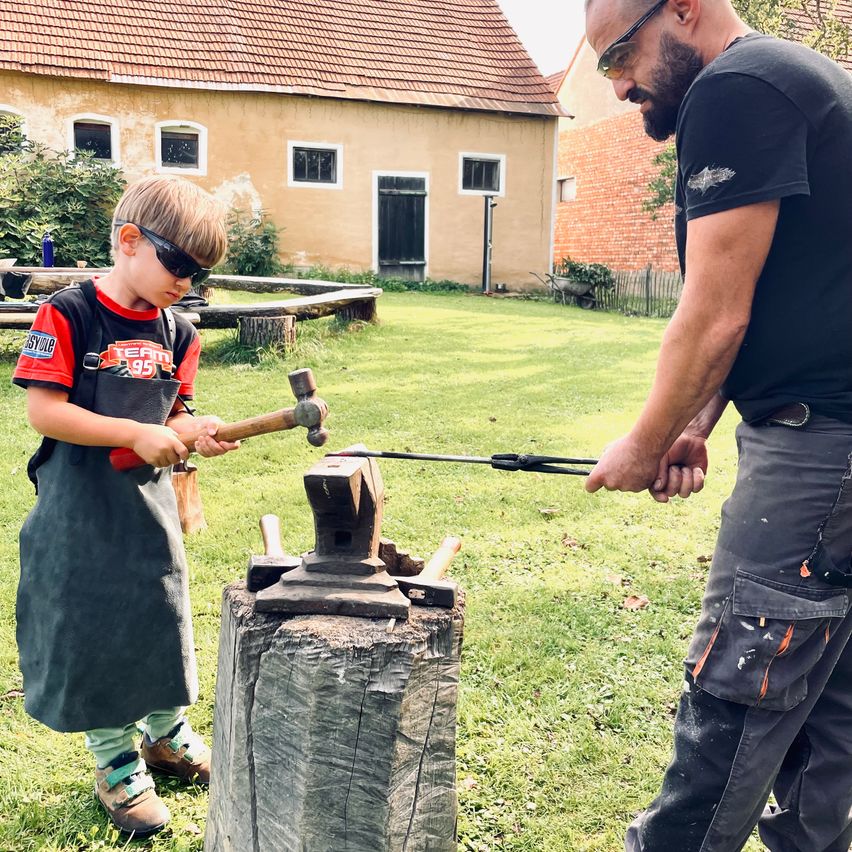 Ein junger Junge in einem roten Shirt und mit Schutzbrille schlägt mit einem Mann in der Nähe auf einem Amboss ein Metallobjekt.