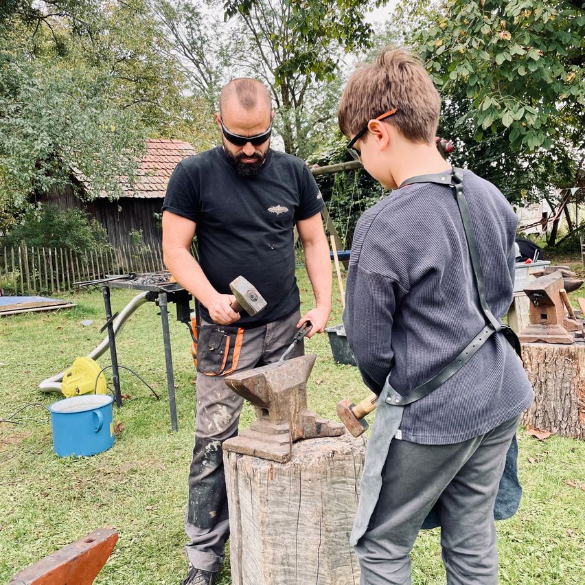 Ein Mann und ein Junge arbeiten im Freien an einem Metall Amboss. Der Mann hält einen Hammer und der Junge beobachtet. Hinter ihnen steht ein Holzhaus und ein Zaun.