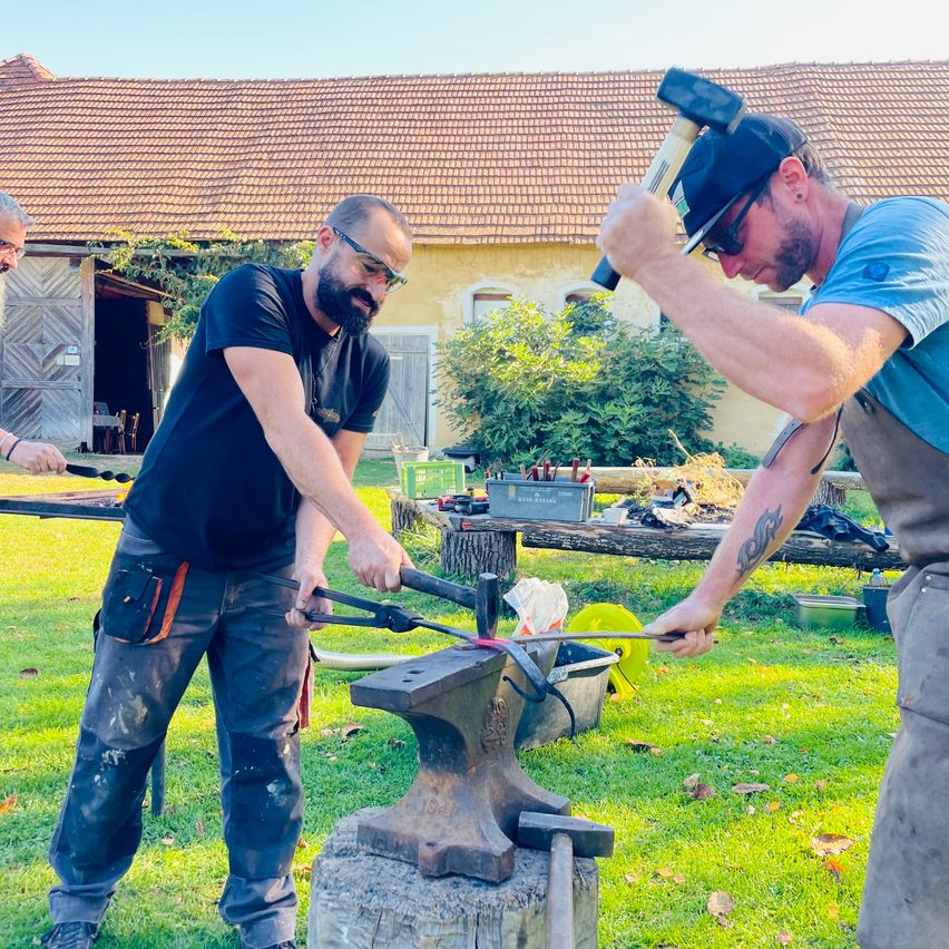 Drei Männer arbeiten draußen an einem Metall Amboss. Ein Mann hält einen Hammer und ein anderer hält eine Zange.