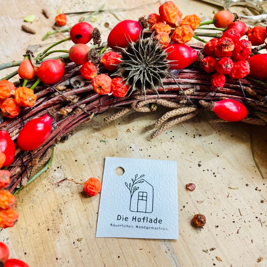 Ein Kranz aus Zweigen und roten Beeren liegt auf einer Holzoberfläche. Ein kleines weißes Schild mit dem Text 'Die Hoflade' und einer Hausillustration ist am Kranz befestigt.