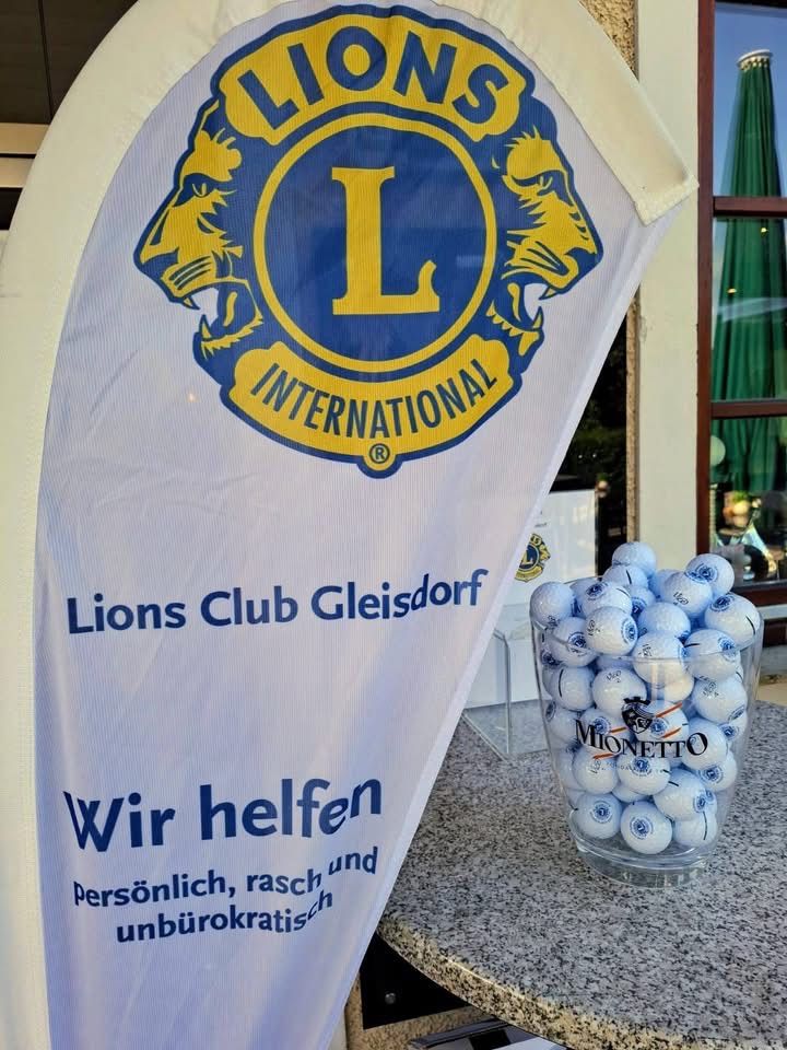 Ein weißes Banner mit dem Logo des Lions Club Gleisdorf und dem Slogan 'Wir helfen'. Daneben eine Glasschale mit Golfbällen.