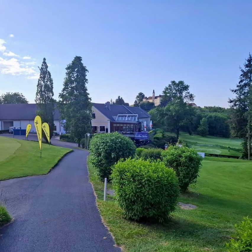 Ein klarer Tag auf einem Golfplatz. Ein Weg führt zu einem Haus mit einem blauen Schild. Gelbe Fahnen stehen im Gras, und ein blauer Müllcontainer ist sichtbar. Bäume und Büsche sind in der Umgebung.