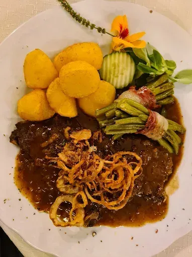 Ein Teller mit gebratenem Rindfleisch in brauner Sauce, gebratenen Zwiebeln, Kartoffelpüree und grünen Bohnen mit Speck.