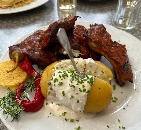 Ein weißer Teller enthält eine gebackene Kartoffel mit Sauce, einen Löffel und Rippchen. Andere Gerichte umfassen Pasta und Getränke.