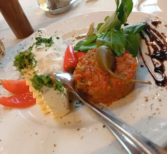 Ein weißer Teller mit einem Fleischbällchengericht, überzogen mit Grünzeug, einer roten Sauce und Tomatenscheiben. Ein Löffel liegt auf dem Teller.
