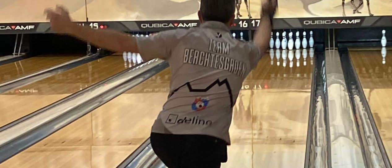 Ein Mann in einem Bowling-Trikot rollt einen Bowling-Ball die Bahn hinunter. Hinter ihm ist eine Wand mit Kamelen und Bowling-Pins.
