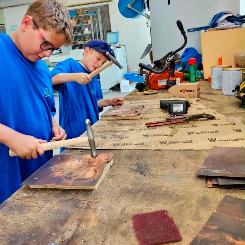 Two boys in blue shirts and hats are hammering metal sheets on a workbench, surrounded by tools and materials.