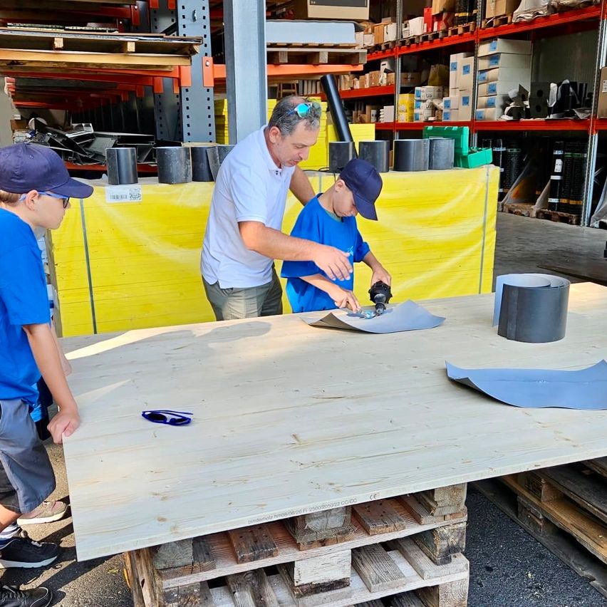An adult man and two boys are working on a wooden table in a warehouse. The man is holding a tool, and the boys are looking at the table.