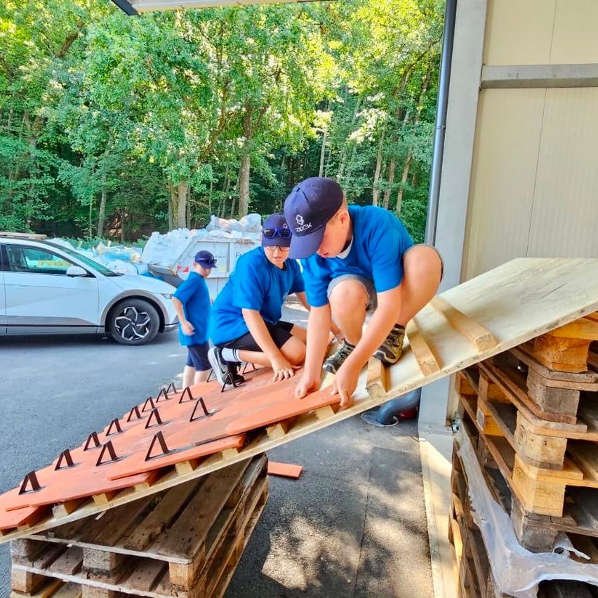 Three boys in blue shirts and caps are working on a wooden structure, possibly a roof, with a car and trees in the background.