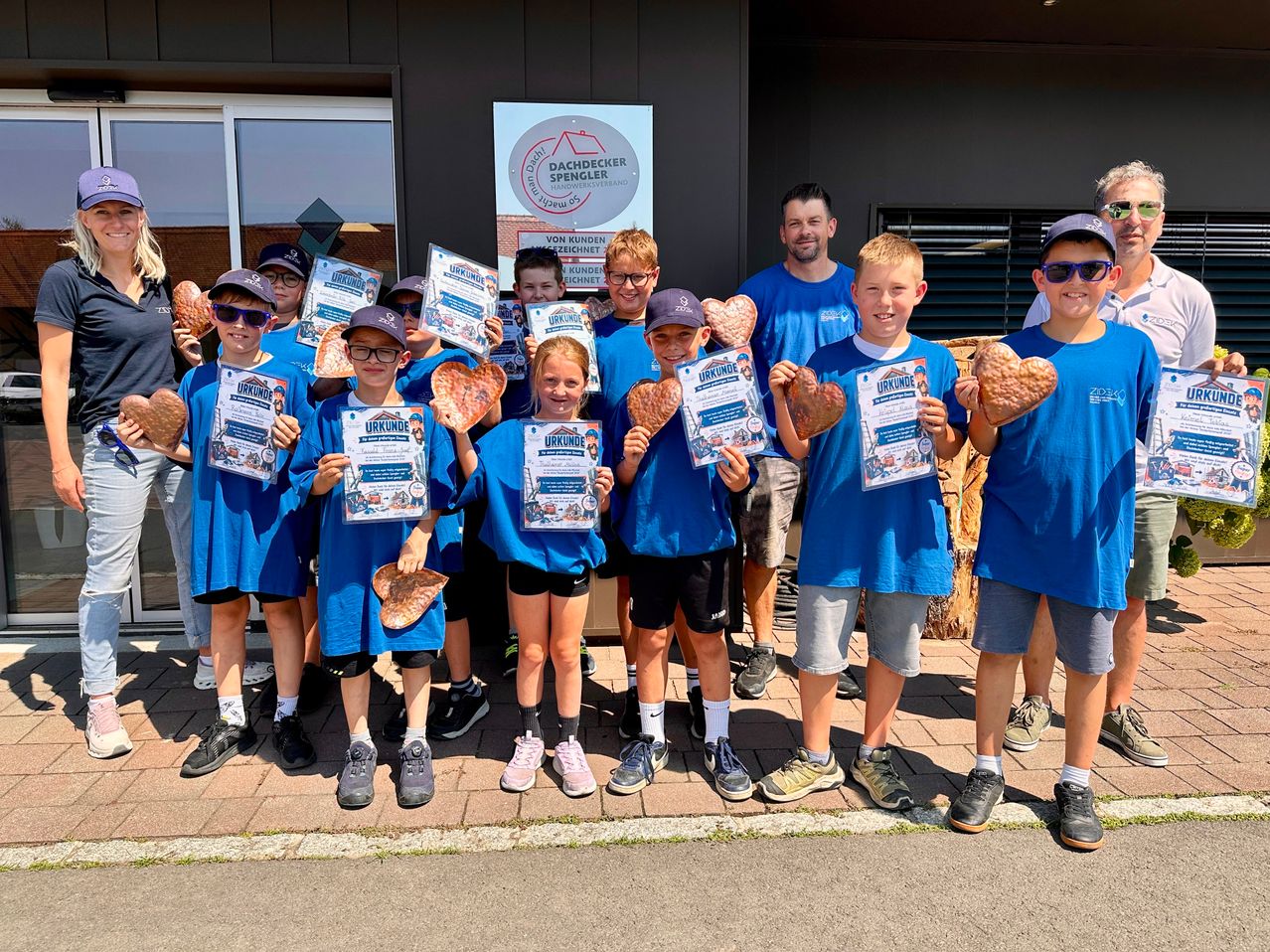 A group of children in blue shirts holding heart-shaped awards, posing for a photo in front of a building.