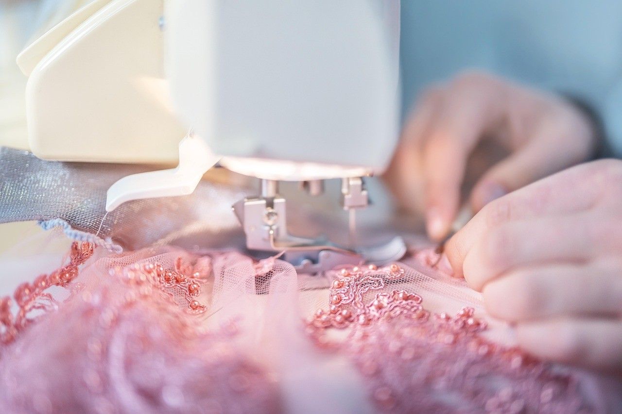 Die Hände einer Person sind dabei zu sehen, wie sie mit einer Nähmaschine an einem rosa Stoff arbeiten. Der Stoff ist zart und mit Perlen und Spitze verziert.