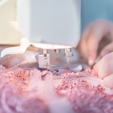 Die Hände einer Person sind dabei zu sehen, wie sie mit einer Nähmaschine an einem rosa Stoff arbeiten. Der Stoff ist zart und mit Perlen und Spitze verziert.