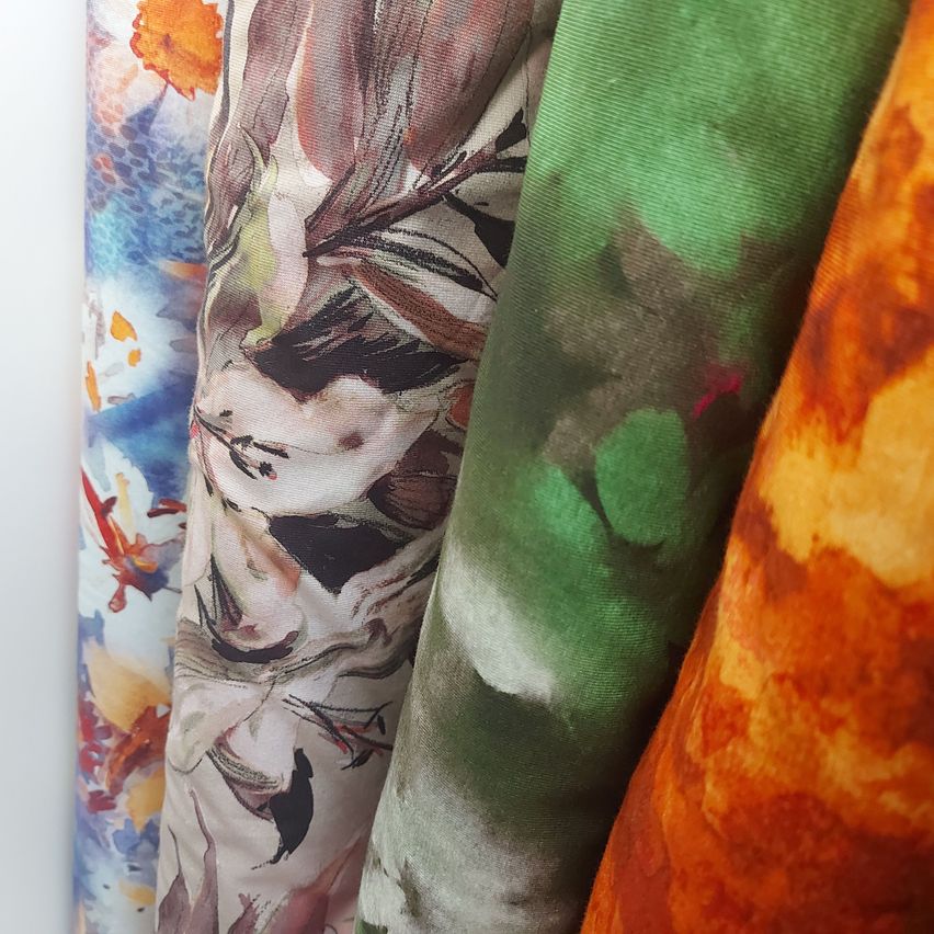 A close-up view of three fabric samples with different abstract patterns. The left fabric has a blue and white watercolor effect. The middle fabric features a brown and black floral design. The right fabric showcases a green and orange abstract pattern.