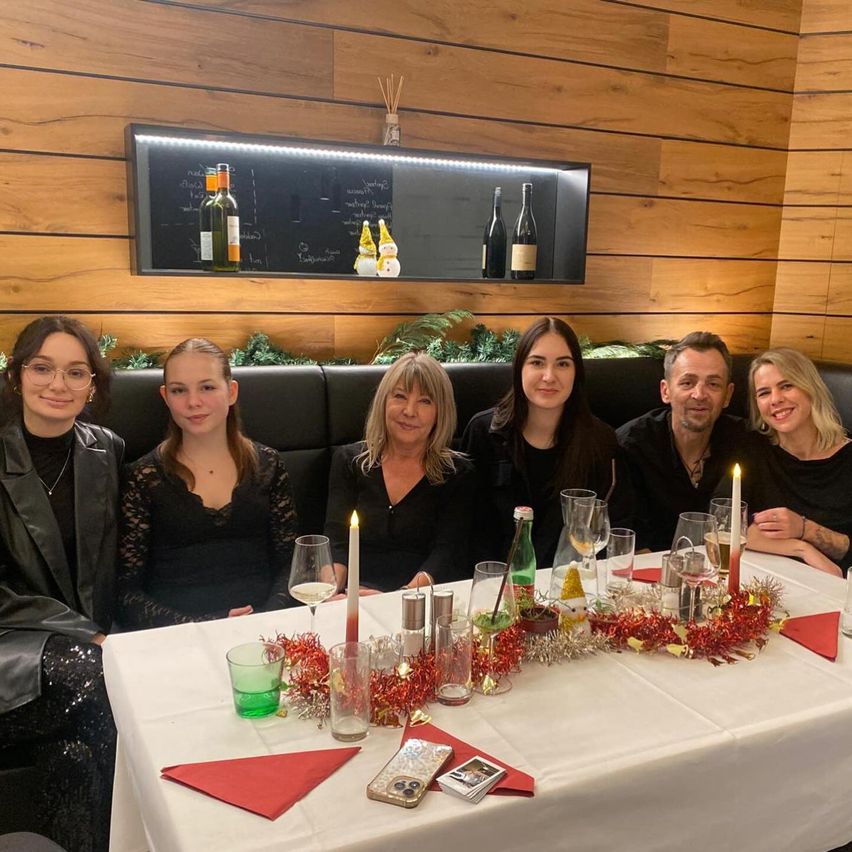 A group of people are sitting around a table in a restaurant. The table is decorated with a Christmas garland, candles, and wine glasses. They are smiling for a photo. Behind them is a wooden wall with a blackboard and wine bottles.