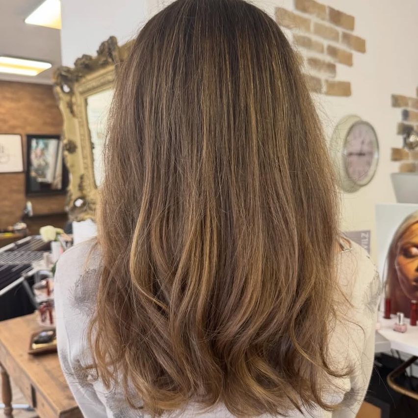 Die Rückseite einer Frau mit langen, welligen braunen Haaren, die in einem Salon steht. Hinter ihr sind ein goldener Spiegel und eine Ziegelwand zu sehen.
