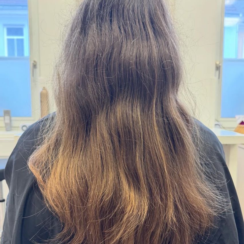 Eine Frau mit langen, welligen, braunen Haaren ist von hinten zu sehen. Das Haar ist gut beleuchtet und zeigt hellbraune Highlights. Der Hintergrund besteht aus einer weißen Wand und zwei Fenstern.