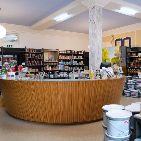 Ein Ladeninneres mit einem großen hölzernen Tresen und Regalen voller Produkte. Der Tresen hat eine Glasplatte. Die Decke hat weißen Marmor und Lichter. Mehrere Farbdosen stehen auf dem Boden.