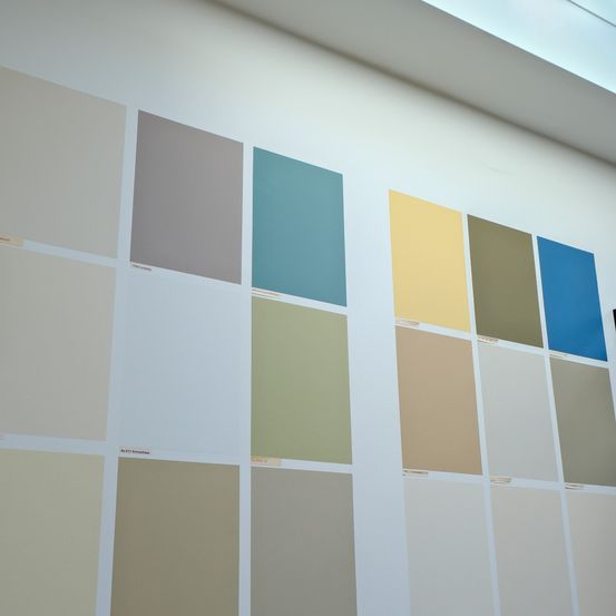 Eine Wand zeigt eine Reihe von Farbmustern, die in einem Raster angeordnet sind, mit verschiedenen Grau-, Blau-, Grün-, Gelb- und Beigetönen.