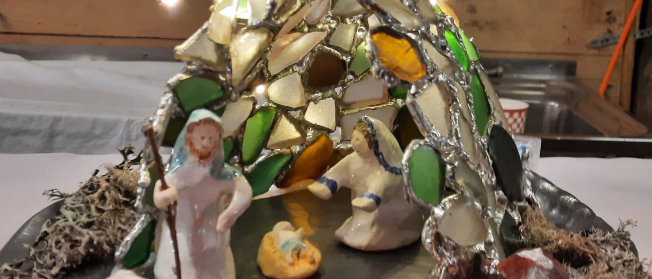 Eine handgefertigte Krippe mit Glasmalereien, mit Maria, Josef, Baby Jesus, einem Schaf und einer Kuh. Eine orange Kerze ist vorn angezündet.