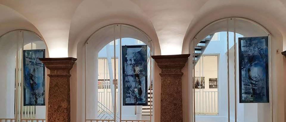 Eine Innenansicht eines Museums mit zwei großen Säulen und drei blau-weißen Gemälden, die zwischen ihnen hängen. Der Raum hat weiße Wände und eine Treppe auf der rechten Seite.