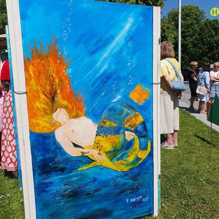 Ein Wandbild einer Meerjungfrau mit orangefarbenen Haaren und einem gelben Schwanz ist auf einem weißen Brett in einem Park ausgestellt. In der Nähe stehen Leute, einige schauen zu.