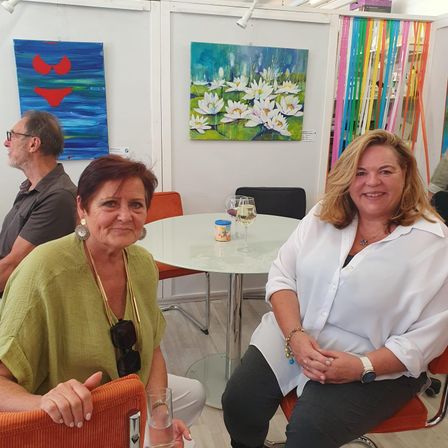 Drei Personen sitzen in einer Kunstgalerie, zwei Frauen lächeln. Vor ihnen steht ein Tisch mit Weingläsern. Die Galerie hat Gemälde an der Wand und bunte Bänder.