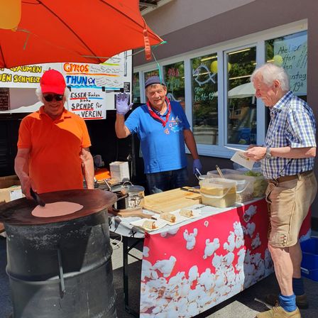 Drei Männer bereiten Essen an einem Außenstand vor. Der Mann in Orange wirft etwas auf einen Grill. Ein Mann in Blau hält ein Messer. Ein anderer Mann liest ein Stück Papier.