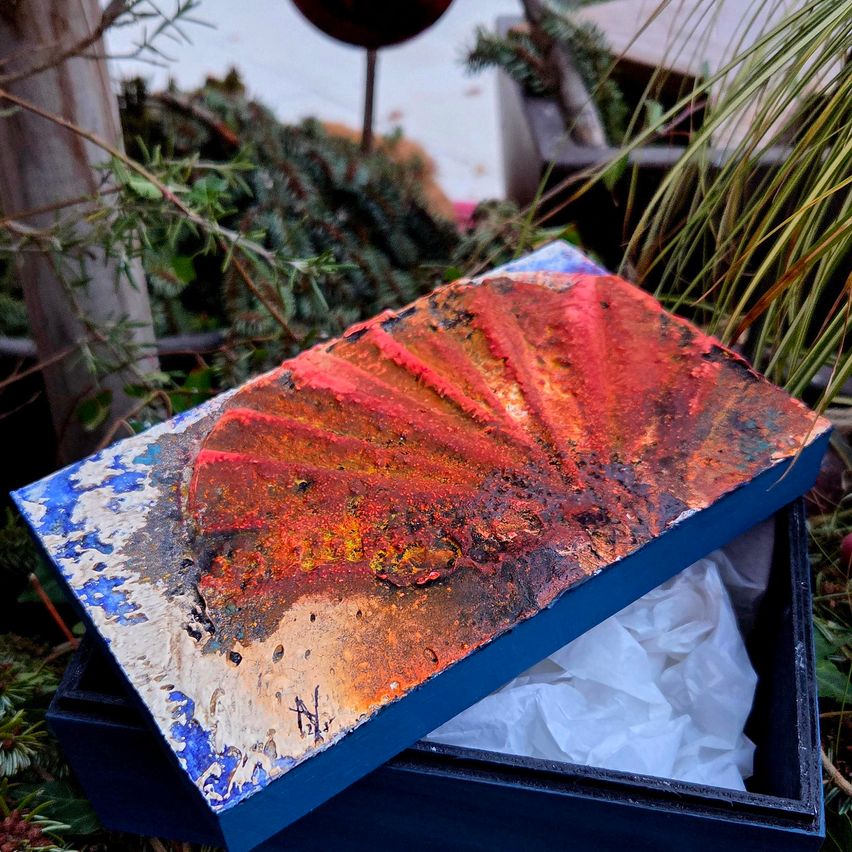 Eine bemalte Muschel auf einer blauen Schachtel mit weißem Papier im Inneren, auf dem Boden mit Pflanzen drumherum platziert.