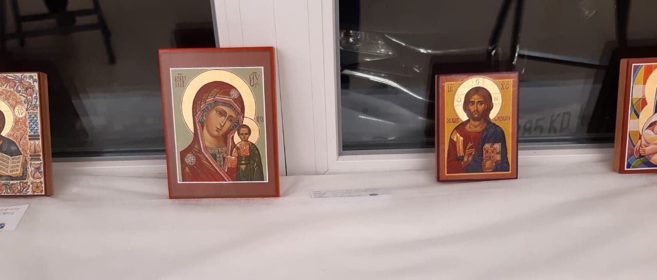 Zwei gerahmte religiöse Ikonen stehen auf einer weißen Oberfläche. Die linke Ikone zeigt eine Frau, die ein Kind hält, beide in traditioneller Kleidung. Die rechte Ikone zeigt einen Mann mit einem Buch. Dahinter spiegelt ein Fenster ein Auto.