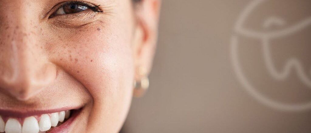 Nahaufnahme des Gesichts einer Frau mit Sommersprossen, die warm lächelt. Ihre Augen sind leicht zusammengekniffen, und sie trägt Ohrringe. Der Hintergrund ist unscharf.