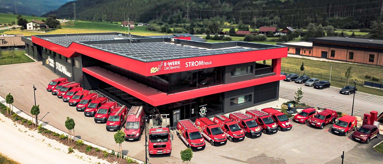 Ein rotes Gebäude mit Solarpanelen auf dem Dach und roten Vans davor, in einer ländlichen Gegend mit Bergen im Hintergrund gelegen.