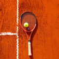Tennisball und Schläger liegen auf rotem Sandplatz mit weißen Linien.