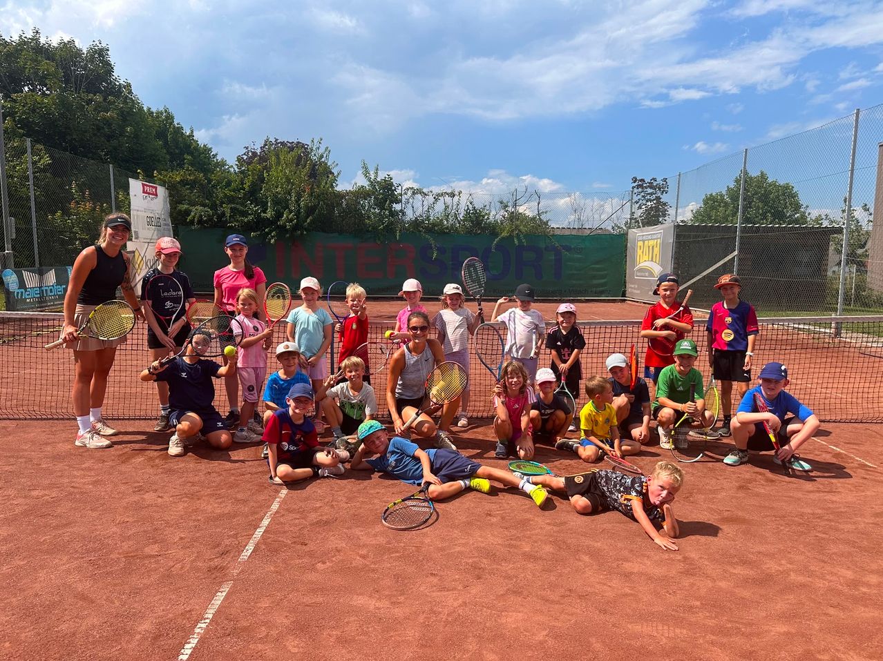Eine Gruppe von Kindern und Erwachsenen posiert für ein Foto auf einem Tennisplatz, hält Tennisschläger und einige tragen Hüte. Der Platz ist von einem Zaun umgeben, auf dem ein Banner mit der Aufschrift INTERSPORT steht.