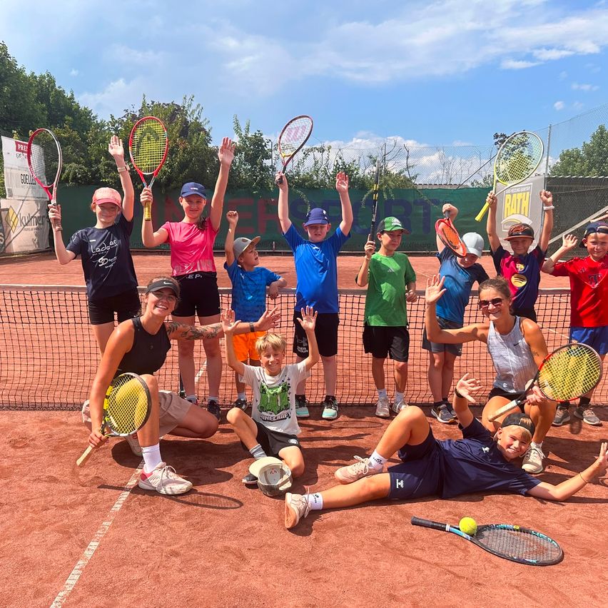 Eine Gruppe von Kindern und Erwachsenen posiert auf einem Tennisplatz für ein Foto, hält Tennisschläger und lächelt.