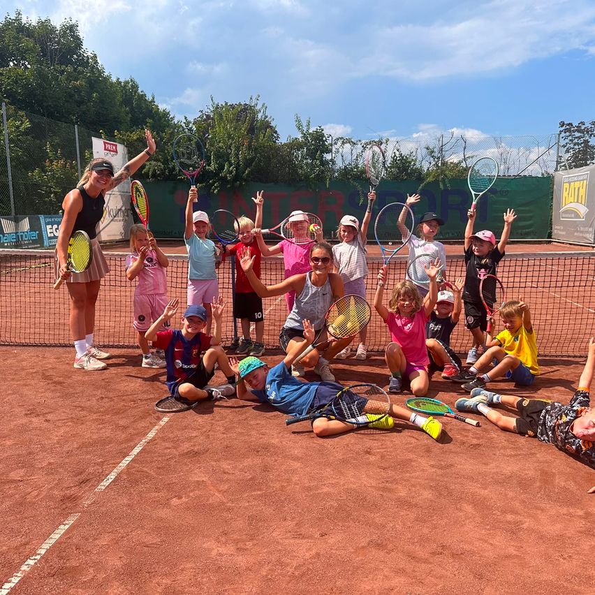 Eine Gruppe von Kindern und einer Frau posiert für ein Foto auf einem Tennisplatz, hält Tennisschläger und lächelt.