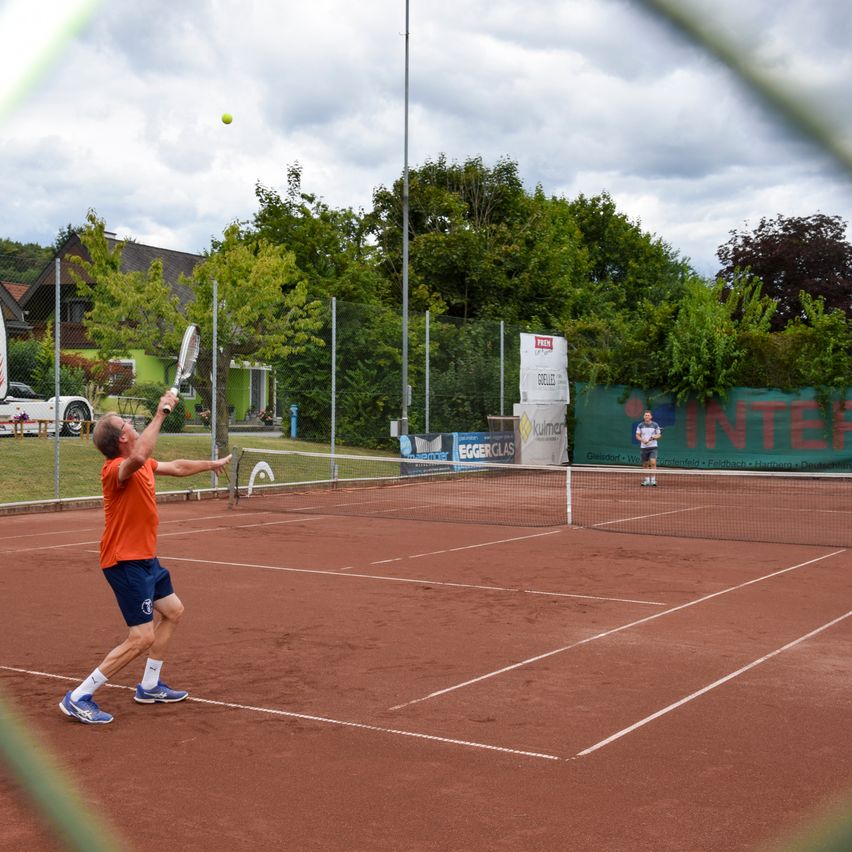 Ein Mann in einem orangefarbenen Shirt und blauen Shorts spielt Tennis auf einem Sandplatz. Er ist dabei, den Ball mit seinem Schläger zu schlagen. Im Hintergrund befindet sich ein Haus und ein Zaun.