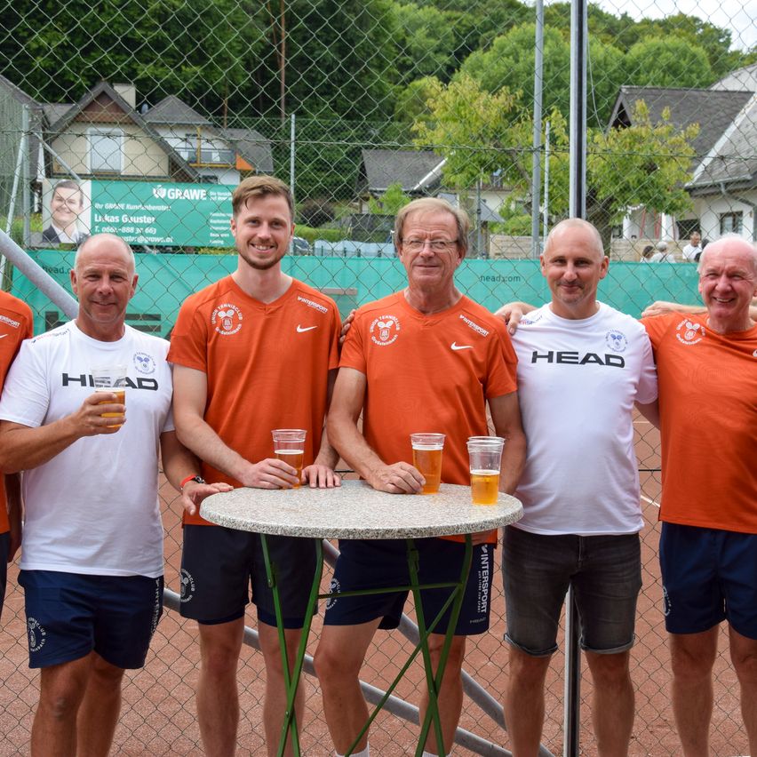 Fünf Männer in orangefarbenen und weißen Trikots posieren für ein Foto vor einem Tennisplatz und halten Getränke. Hinter ihnen steht ein Zaun mit einem Banner und Häusern.